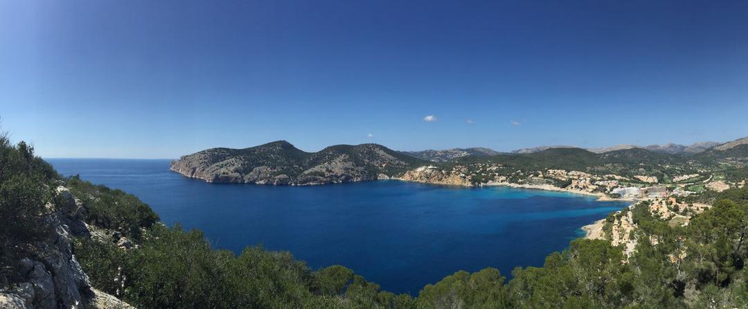 Blick von einer Anhöhe an grün bewachsener Küste der Balearen auf Landzungen und das weite, blaue Meer 